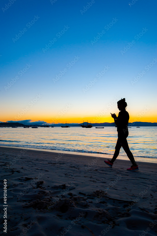 Silhueta de mulher caminhando na praia no fim de tarde mexendo no celular