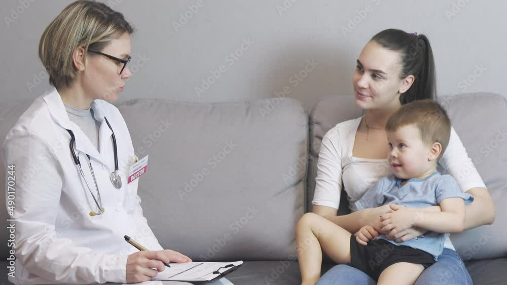 pediatrician doctor visits baby boy child at home. caucasian kid sitting on mother's knees on grey sofa living-room. female woman doctor in white uniform, stethoscope, talking parent asking symptoms pediatrician doctor visits baby boy child at home. caucasian kid sitting on mother's knees on grey sofa living-room. female woman doctor in white uniform, stethoscope, talking parent asking symptoms