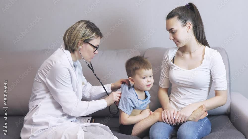 pediatrician doctor visits baby boy child at home. caucasian kid and mother sitting on grey sofa living-room. female woman doctor in white uniform, glasses, listening toddle's back using stethoscope, pediatrician doctor visits baby boy child at home. caucasian kid and mother sitting on grey sofa living-room. female woman doctor in white uniform, glasses, listening toddle's back using stethoscope,