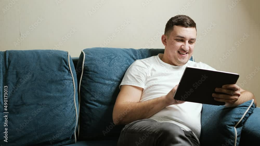 A 30-year-old man of European appearance sits on a dark blue couch and works at a tablet. Guy smiles looking at the tablet screen. A 30-year-old man of European appearance sits on a dark blue couch and works at a tablet. Guy smiles looking at the tablet screen.