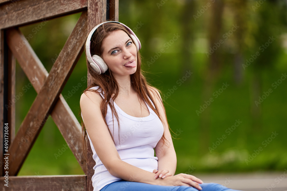 Woman in wireless headphones shows tongue in public park. Woman in wireless headphones shows tongue in public park.