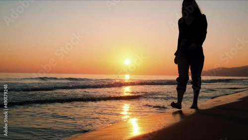 Slow motion silhouette woman feet walking barefoot by beach at golden sunset footprints in sand. Female tourist on summer vacation. Woman in beautiful waving dress at sunset