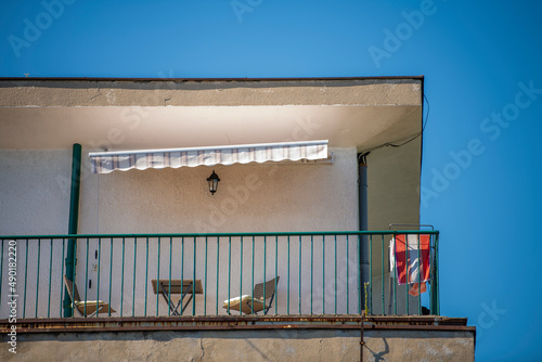 Old Vintage balcony or terrace in a multi story residential building, with desk and chairs, laundry dryer and colorful awning. Hot summer day. Blue sky in background. photo