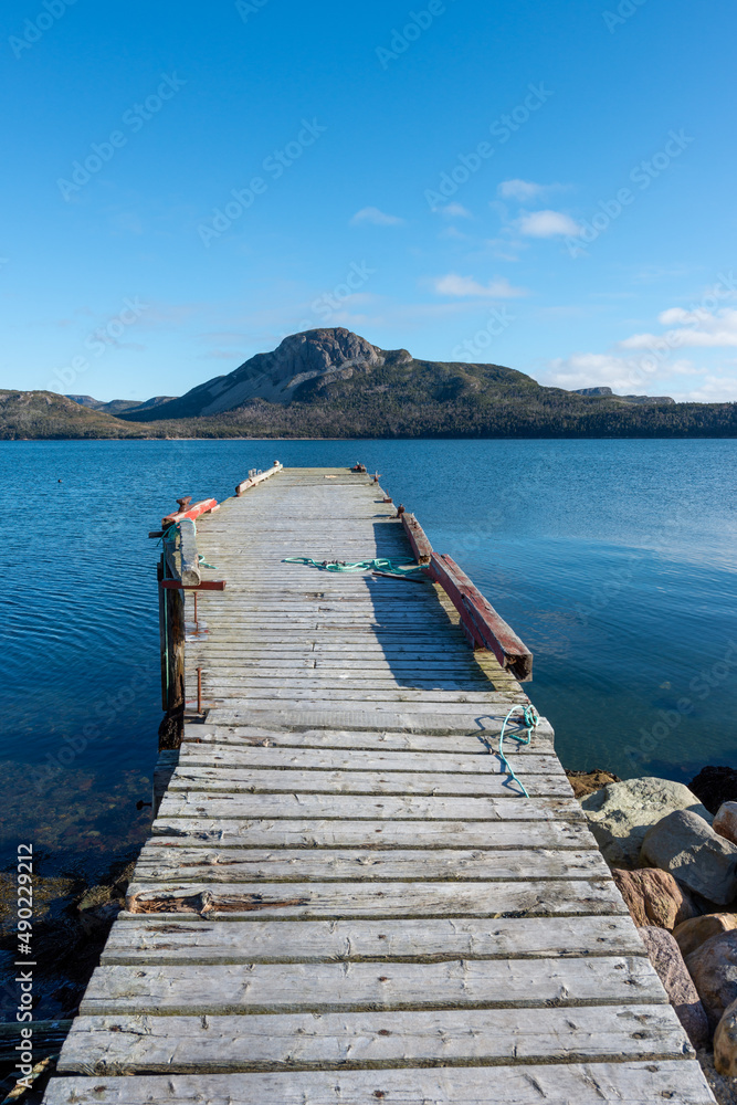 An old wooden pier with a yellow color mooring deck. The wharf juts out into the calm ocean with tree covered mountains in the bay or cove. The sky and water are clear deep blue colors. 