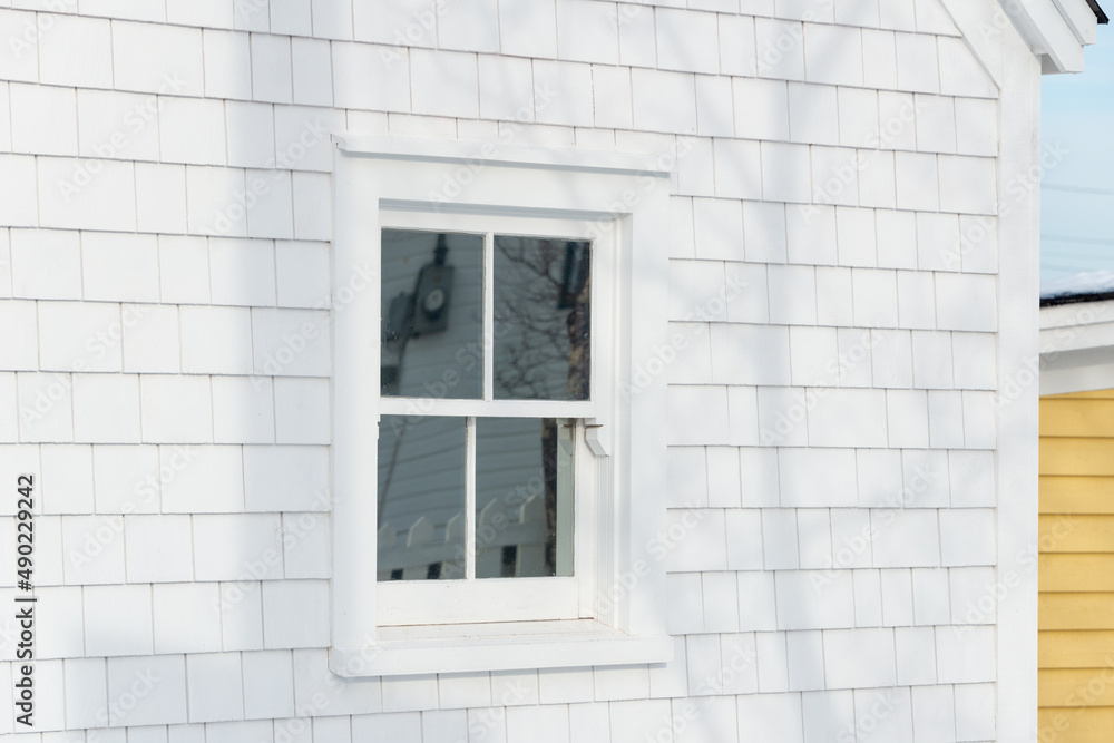 Fototapeta premium The exterior corner of a vintage wooden building with a closed glass single hung window. The wall is covered in white shingles. There's a yellow wooden building in the background with white trim. 