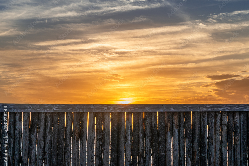 A wooden fence made from logs lined up in front of a vibrant ocean. The seawall is wood with narrow trees cut the same height. The background is a vibrant orange evening sky with whispy clouds.