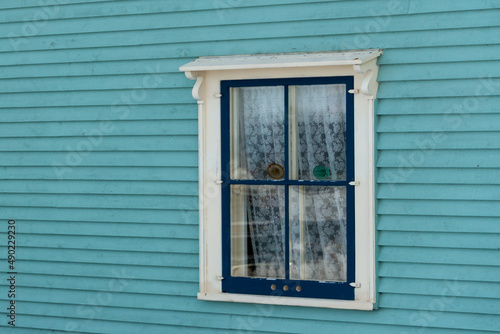 The exterior outside wall of a teal blue heritage wood building with dark green and tan trim. The country style house has narrow clapboard and a single double hung window with lace curtains hanging. 