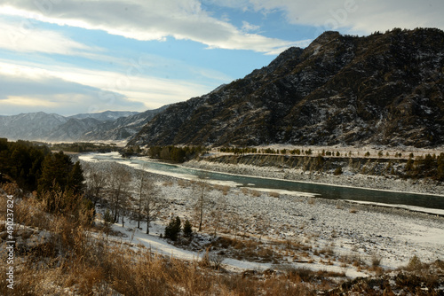 Snow-covered banks of a beautiful river flowing through a picturesque valley at the foot of high mountains in early winter.