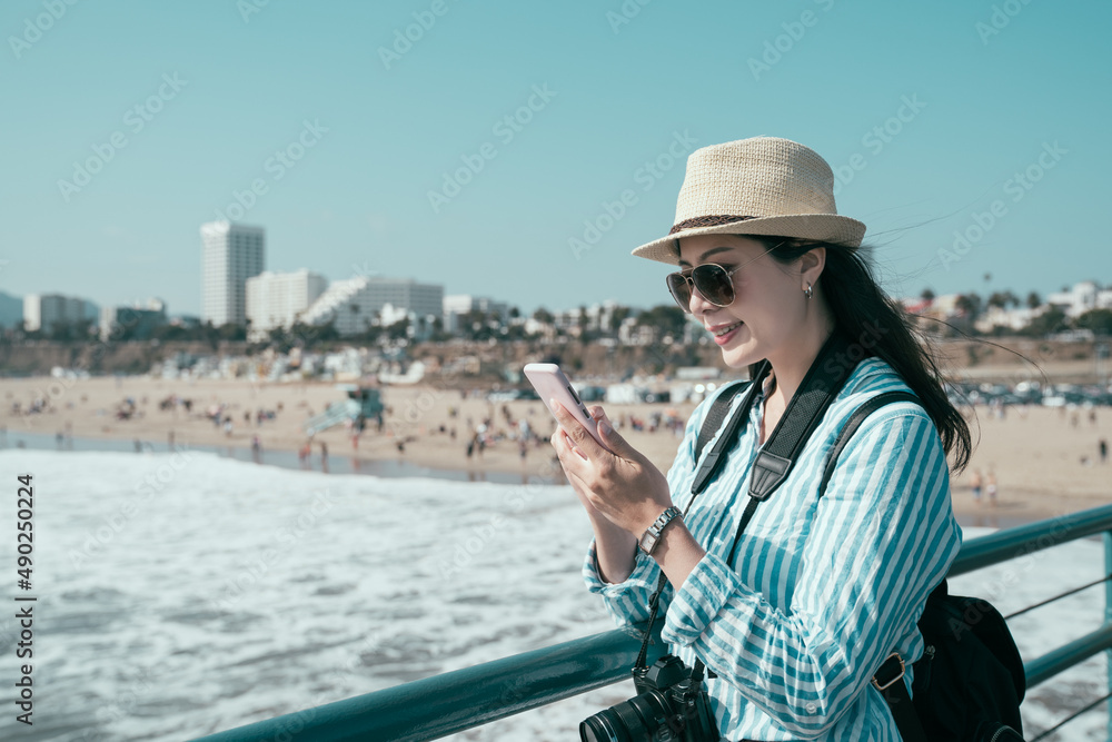 Happy young asian chinese girl backpacker looking mobile phone screen in beautiful summer sunny day near sea port. cheerful woman travel photographer holding cellphone and leaning on railing.