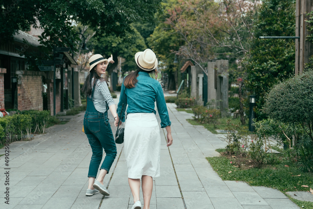 Φωτογραφία back view full length of young excited girl in denim overalls walking and hands holding friend on street