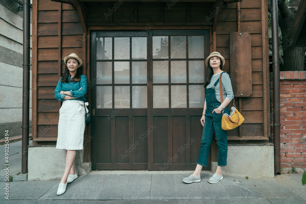 full length front view of two asian girl travelers standing by wooden door of traditional japanese ancient house. charming women tourists leaning on wood wall with copy space outdoor vintage style. full length front view of two asian girl travelers standing by wooden door of traditional japanese ancient house. charming women tourists leaning on wood wall with copy space outdoor vintage style.