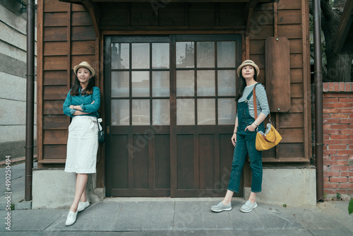 full length front view of two asian girl travelers standing by wooden door of traditional japanese ancient house. charming women tourists leaning on wood wall with copy space outdoor vintage style.