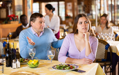 Smiling man trying to calm his upset girlfriend offended after disagreement or quarrel while sitting at table in cozy restaurant during dinner