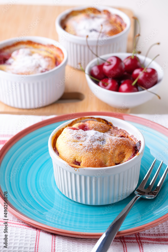 Traditional french baked cherry pie clafoutis served in white ramekin and some cherry on the table. Vertical. Traditional french baked cherry pie clafoutis served in white ramekin and some cherry on the table. Vertical.