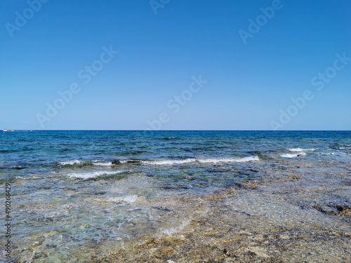 The coast of the Mediterranean Sea, waves, clear water through which the bottom is visible. All this against a blue sky with clouds.