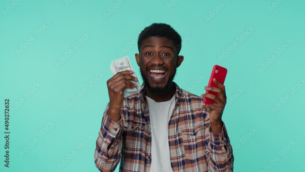 Happy african american man in shirt looking mobile phone display sincerely rejoicing win, receiving money dollar cash banknotes, success lottery luck. Young guy isolated on gray studio background Happy african american man in shirt looking mobile phone display sincerely rejoicing win, receiving money dollar cash banknotes, success lottery luck. Young guy isolated on gray studio background