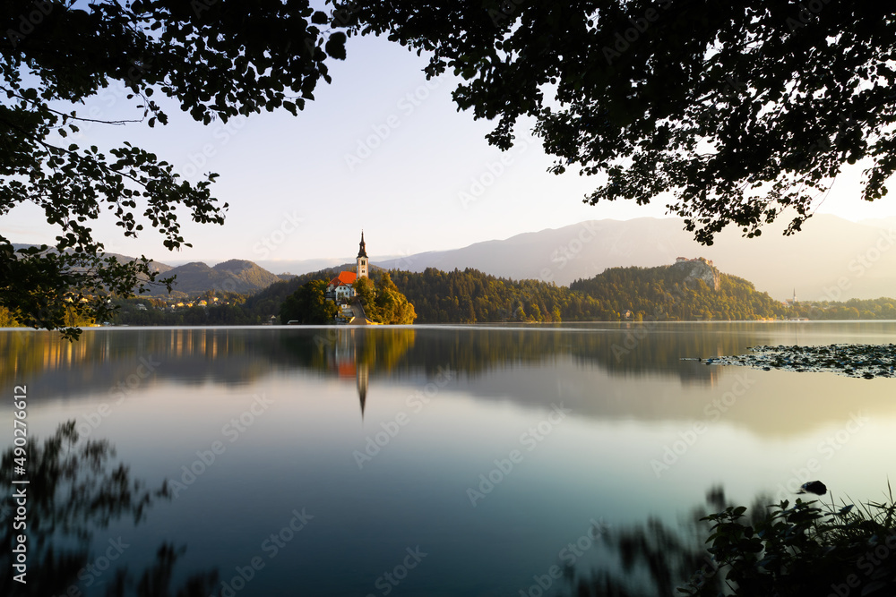 Obraz premium Lake Bled with a church on an island at sunrise through window of branches. Peaceful scenery with a religion building in the middle of nature illuminated with morning light with copy space.