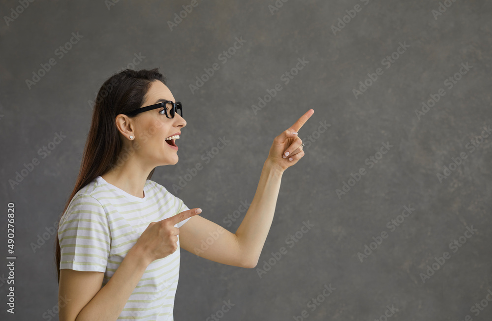 Joyful woman with a pleasantly surprised expression presents a new proposal on a gray background. Profile view of young woman pointing fingers at free space for text or promotional products. Banner. Joyful woman with a pleasantly surprised expression presents a new proposal on a gray background. Profile view of young woman pointing fingers at free space for text or promotional products. Banner.