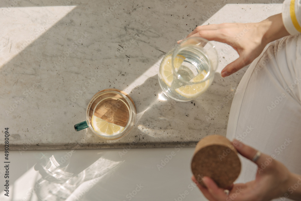 Top view of glass with drink near jug of lemonade in sunlight. Detail of body part of young fair-skinned girl holding wooden cover. Windowsill breakfast concept Top view of glass with drink near jug of lemonade in sunlight. Detail of body part of young fair-skinned girl holding wooden cover. Windowsill breakfast concept