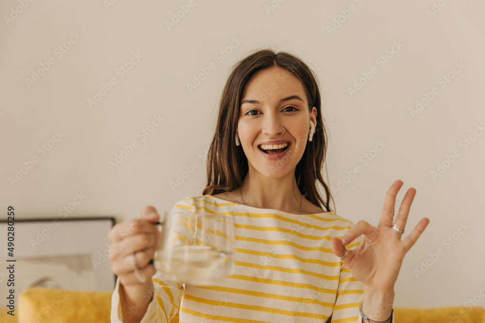 Portrait of happy energetic girl showing okay gesture indoors. Brown-haired woman smiles strongly while looking into camera and holds glass cup in her hands. Relaxation and comfort concept. Portrait of happy energetic girl showing okay gesture indoors. Brown-haired woman smiles strongly while looking into camera and holds glass cup in her hands. Relaxation and comfort concept.
