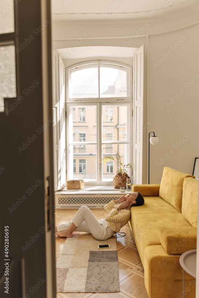 Far away photo of fair-skinned young woman sitting on floor with head leaning against sofa in cozy room. Relaxed brunette listens to music through headphones, wearing yellow blouse and white pants.