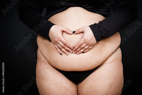 a woman shows a heart with her hands on a fat belly on a black background. obese person. body positivity concept