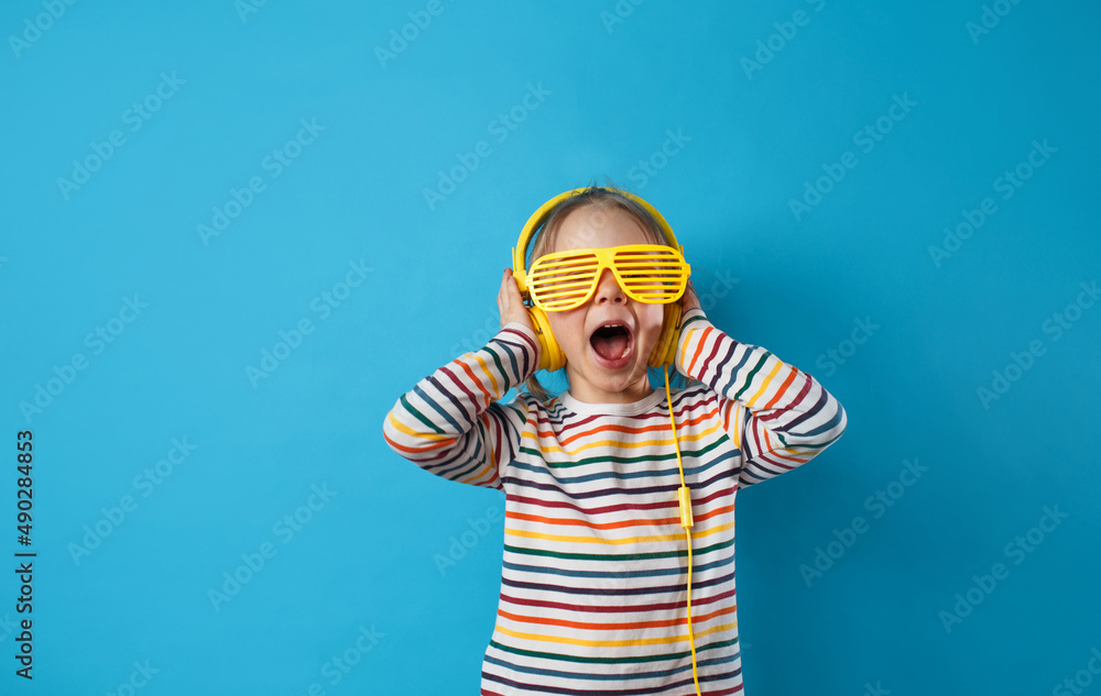 portrait of a cute charming girl listening to her favorite song in yellow headphones and stylish glasses on a blue background