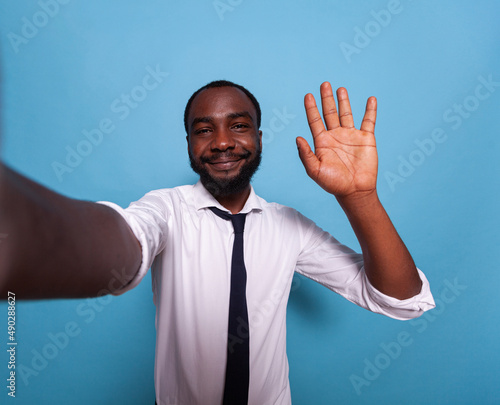 Smiling vlogger waving hello at camera in videocall conference on blue background. Wide angle pov of influencer taking a smartphone selfie doing hand gesture for social media. photo