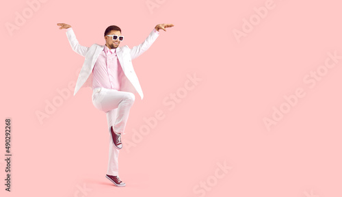 Funny man posing in fashion studio. Full body happy attractive young guy wearing white suit, pink shirt, cool sunglasses and trainers standing on one foot, arms spread apart, on text space background photo