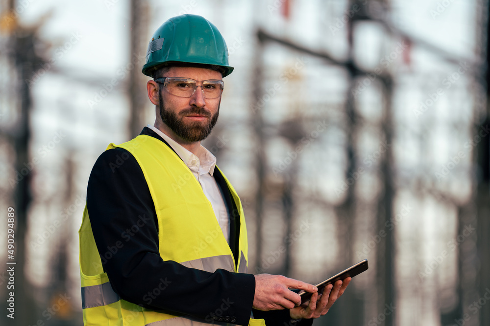 Electric engineer wearing helmet suit and safety vest checking datas on tablet with substation in background Electric engineer wearing helmet suit and safety vest checking datas on tablet with substation in background