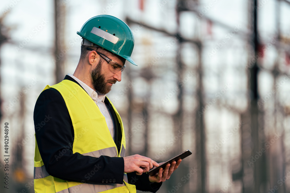 Obraz premium Electric engineer wearing helmet suit and safety vest checking datas on tablet with substation in background