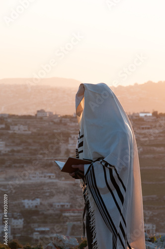 A Jewish man holding a siddur prayerbook and wearing a tallit and tefillin prays the morning service at sunrise on a hilltop in the Judean Mountains of Israel photo