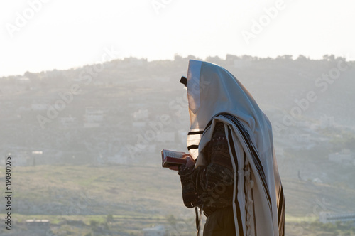 A Jewish man holding a siddur prayerbook and wearing a tallit and tefillin prays the morning service at sunrise on a hilltop in the Judean Mountains of Israel