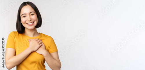 Romantic asian girfriend, holding hands on heart, smiling with care and tenderness, standing in yellow tshirt over white background photo