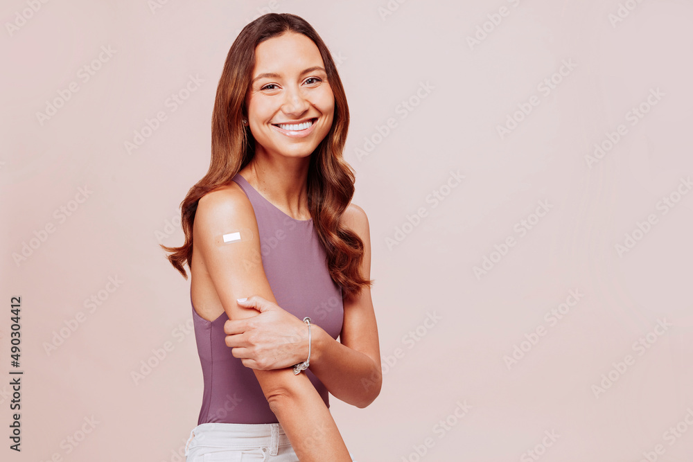 Portrait of cheerful young mixed race vaccinated woman showing bandaged arm at studio over beige background. Female after receiving vaccination. Copy space. Portrait of cheerful young mixed race vaccinated woman showing bandaged arm at studio over beige background. Female after receiving vaccination. Copy space.