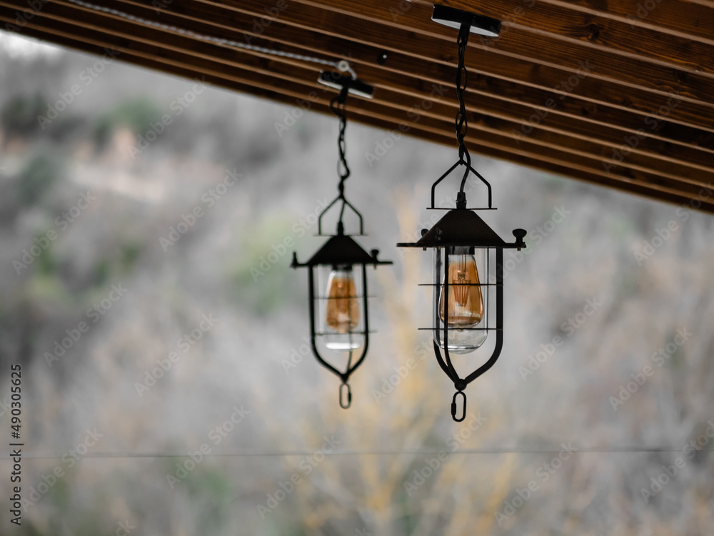 Two lanterns with large orange bulbs hang from the wooden porch of a rural town house and the forest vegetation out of focus in the background Two lanterns with large orange bulbs hang from the wooden porch of a rural town house and the forest vegetation out of focus in the background