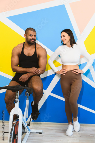 Athletic couple: adult afro american man and young adult caucasian woman take a break and talking near the cycling machine in a sports club.