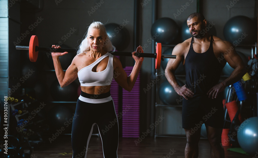 Fit young caucasian woman lifting barbell, working out in a gym with afro-american personal trainer assistance cheering her on. Personal training and bodybuilding concept.