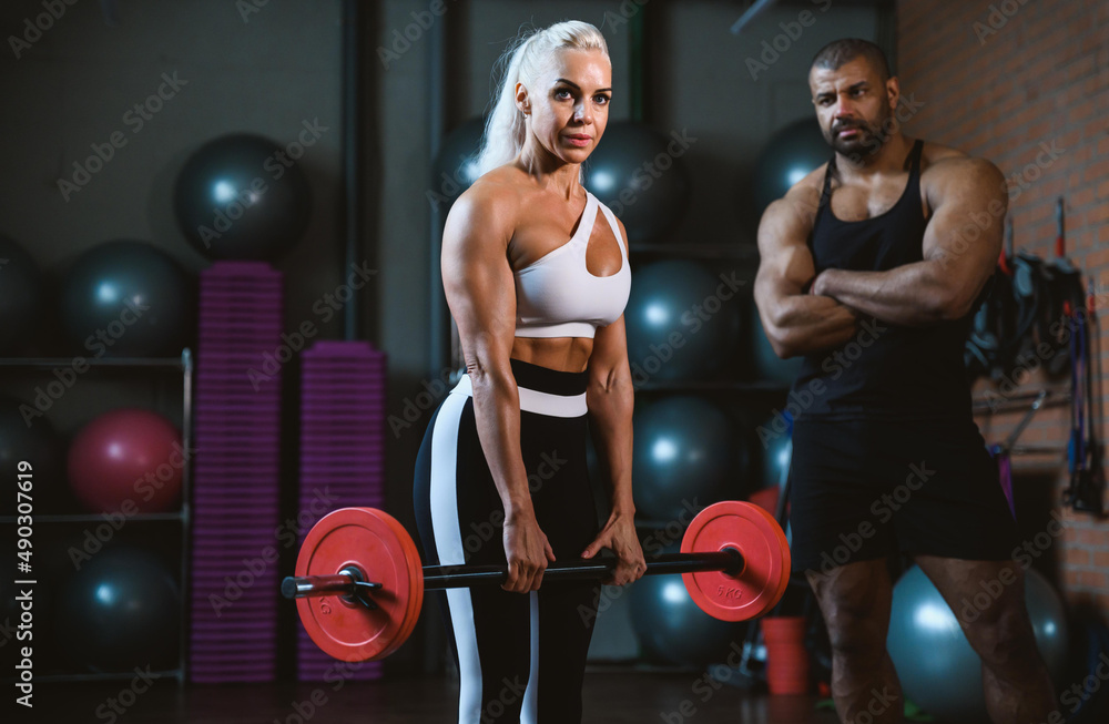 Fit young caucasian woman lifting barbell, working out in a gym with afro-american personal trainer assistance cheering her on. Personal training and bodybuilding concept. Fit young caucasian woman lifting barbell, working out in a gym with afro-american personal trainer assistance cheering her on. Personal training and bodybuilding concept.