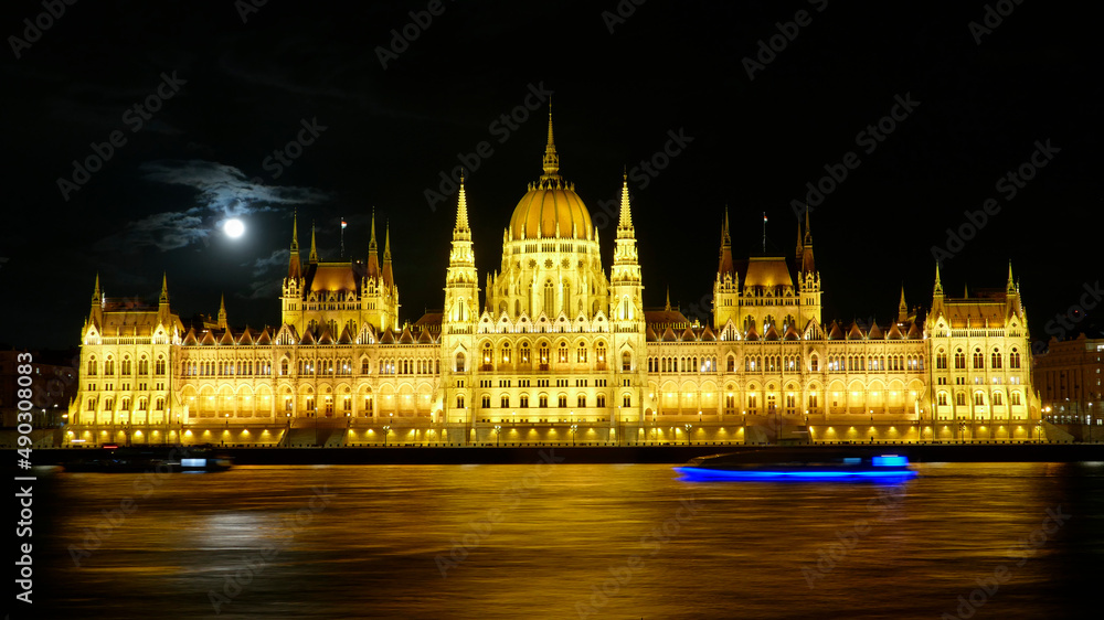 Illuminated Parliament building in Budapest, Hungary by night with full moon and a blue ship on the Danube