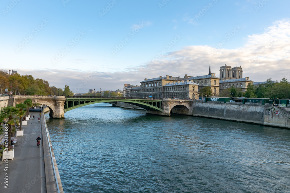 Paris. Pont Notre-Dame bridge. From Pont Au Change bridge. Hôtel-Dieu hospital, Cathedral Notre-Dame de Paris. People walking and cycling along Rives de Seine Park. Seine River water. Blue cloudy sky.