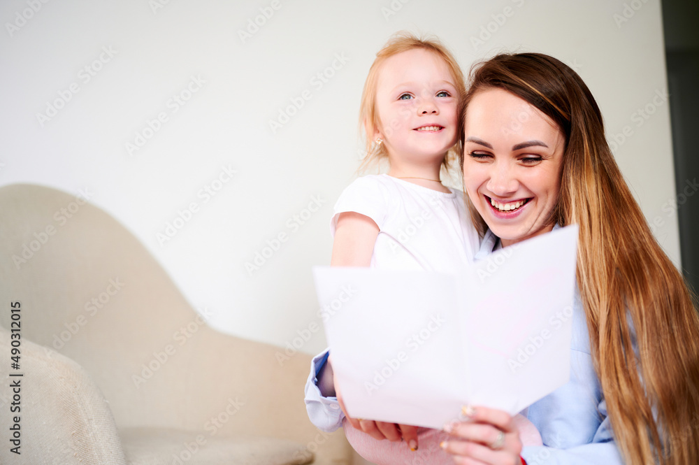 Happy Mother's Day. Happy smiling mom with her little daughter reading holiday card while sitting on the floor Happy Mother's Day. Happy smiling mom with her little daughter reading holiday card while sitting on the floor
