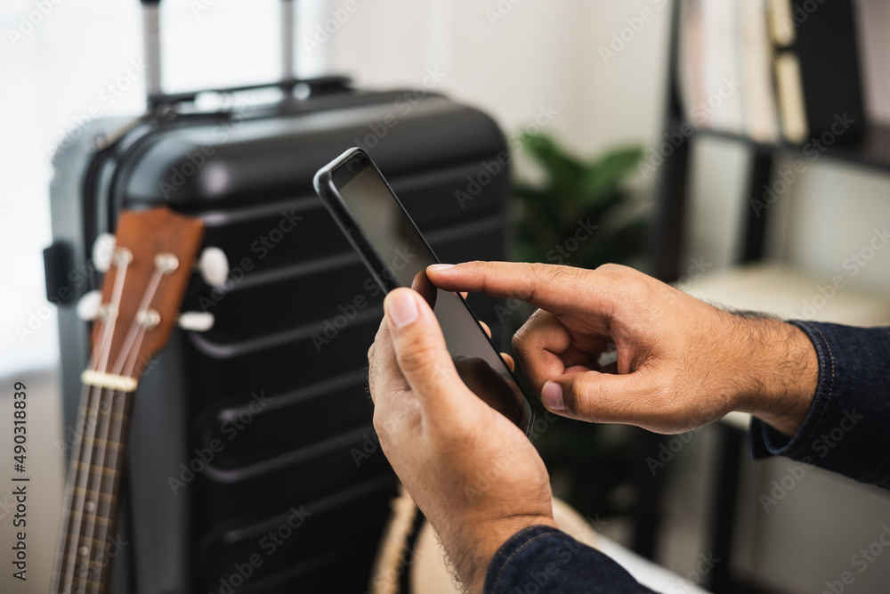 Close up male hands using smartphone near trolley bag in the house. Book a trip on smartphone and pay for it. Search for a route to travel by cell phone