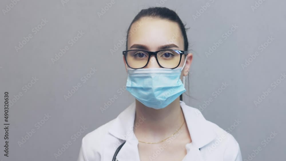 close-up portrait of pretty young woman doctor in white uniform, wearing glasses, facial protective mask, looking to camera. female caucasian professional physician indoors. medicine worker occupation close-up portrait of pretty young woman doctor in white uniform, wearing glasses, facial protective mask, looking to camera. female caucasian professional physician indoors. medicine worker occupation