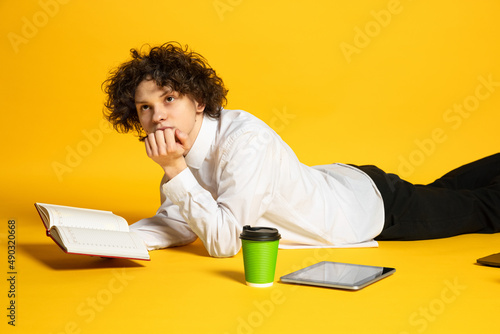 Portrait of young boy, student lying on floor, reading book, doing homework isolated over yellow background. Exams preparation photo