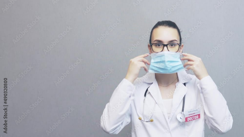pretty young woman doctor in white uniform with stethoscope over neck, wearing glasses puts on facial protective mask, looking to camera. indoors female caucasian professional physician. medicine work pretty young woman doctor in white uniform with stethoscope over neck, wearing glasses puts on facial protective mask, looking to camera. indoors female caucasian professional physician. medicine work