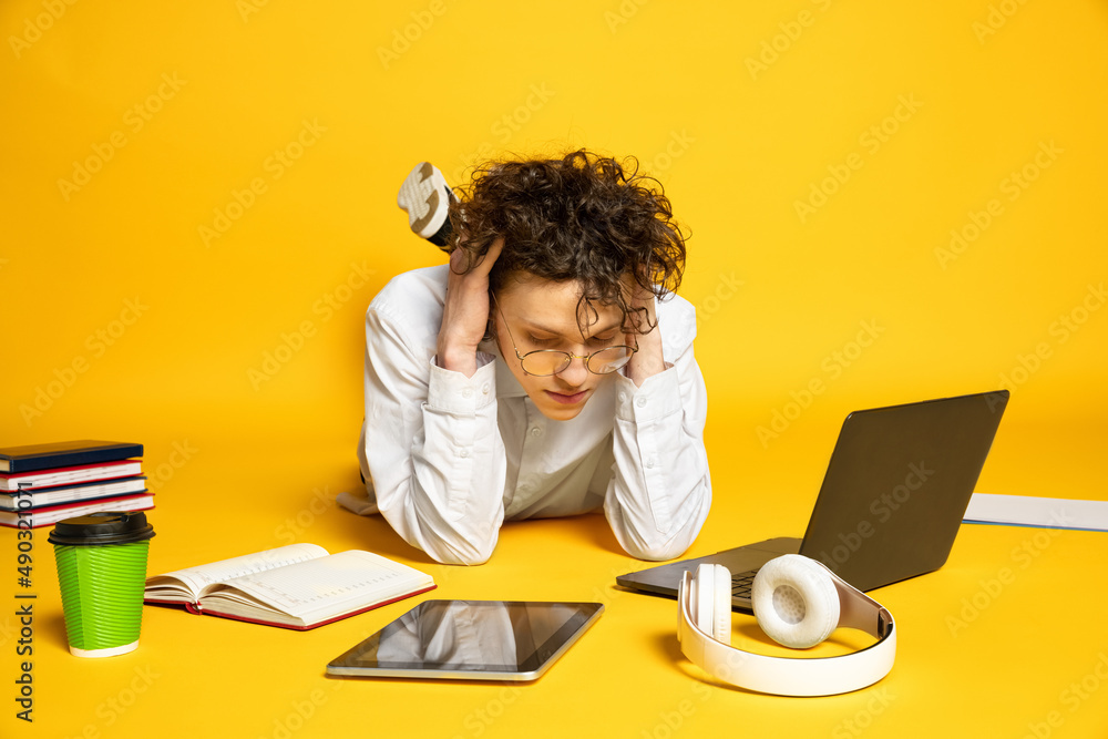 Portrait of young curly man, student in glasses lying on floor around study supplies, studying isolated over yellow studio background Portrait of young curly man, student in glasses lying on floor around study supplies, studying isolated over yellow studio background