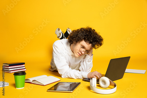 Portrait of young man, studnt studying, making homework on laptop isolated over yellow studio background photo