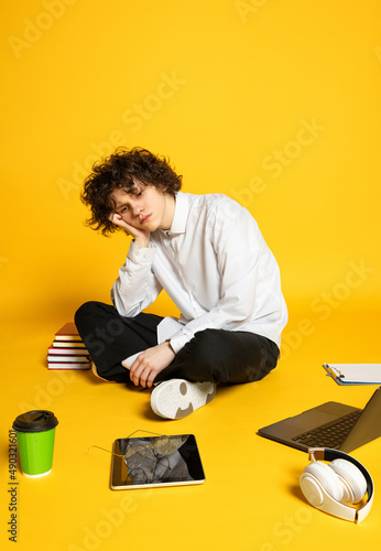 Portrait of young curly man, student sitting on floor arounf study supplies isolated over yellow background photo