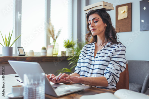 Portrait of a young attractive woman at the desk with books on her head, sitting straight, working on laptop. Education concept photo, lifestyle.